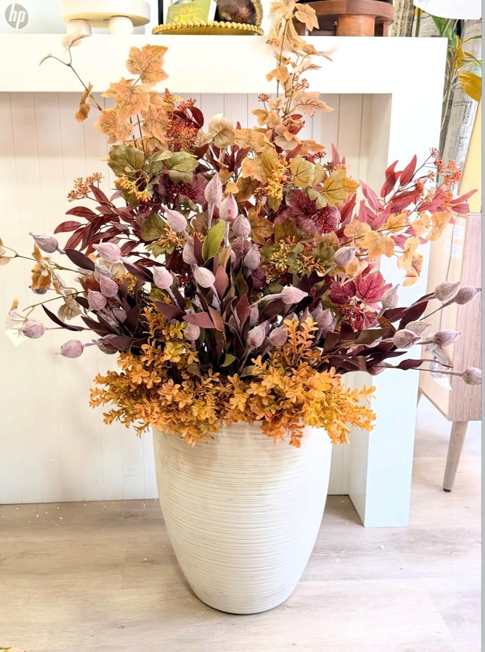 Burgundy, gold and muted green foliage arranged in a tall ribbed ivory planter beside a white counter.