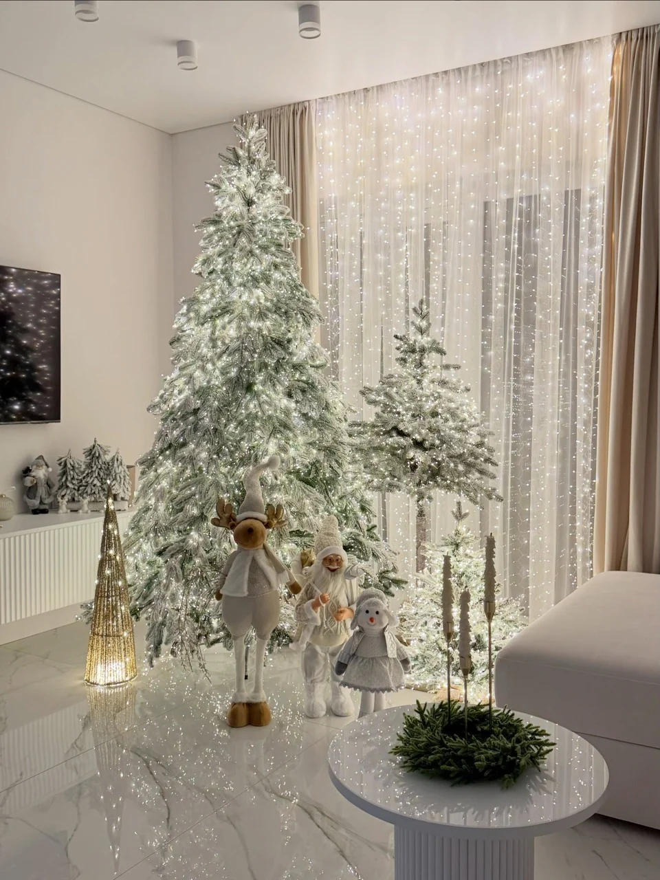 Three illuminated flocked trees with festive figures and curtain lights beside a white sofa and round coffee table.