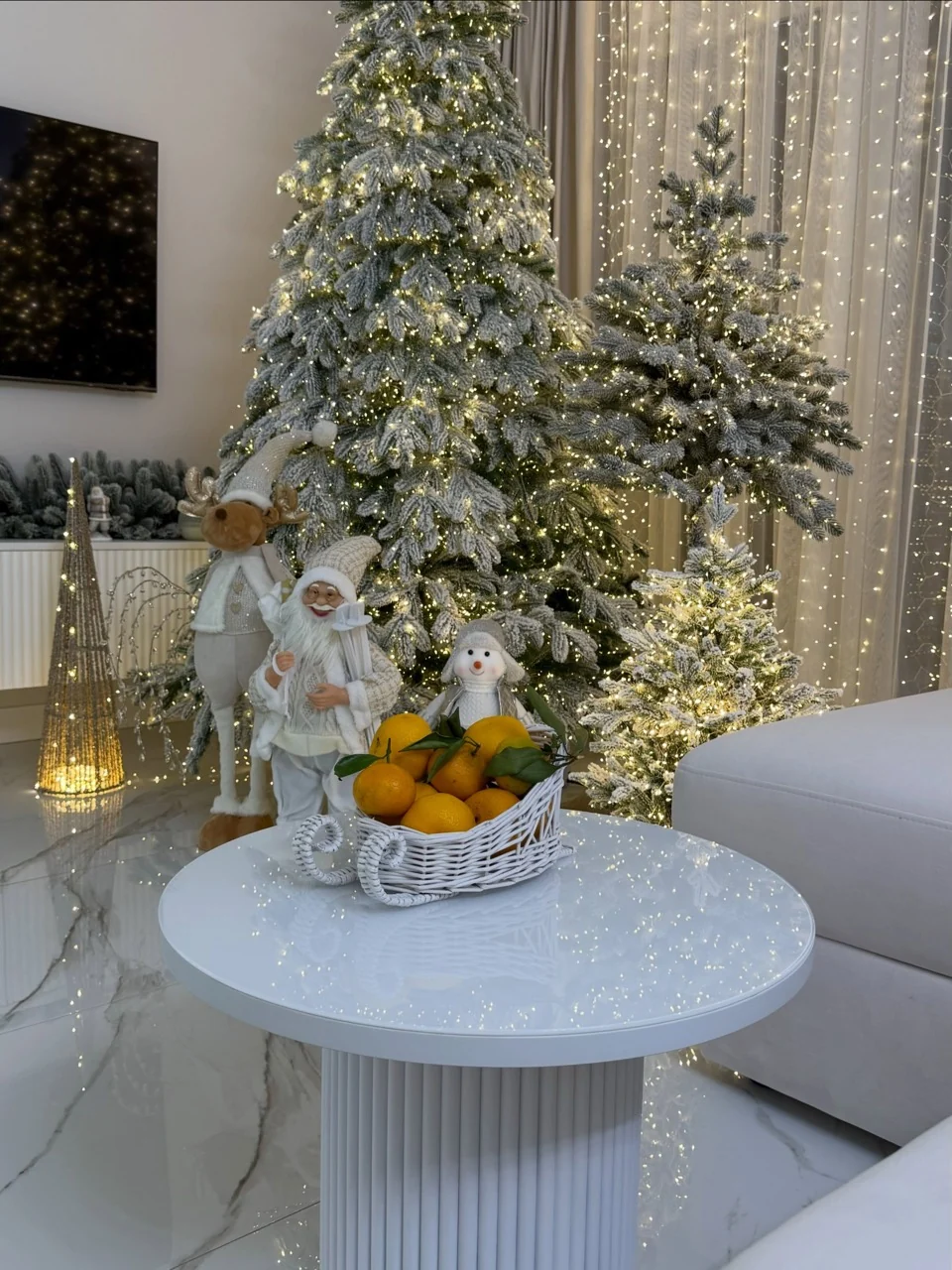 A citrus-filled white basket on a round coffee table in front of illuminated flocked Christmas trees and festive figures.
