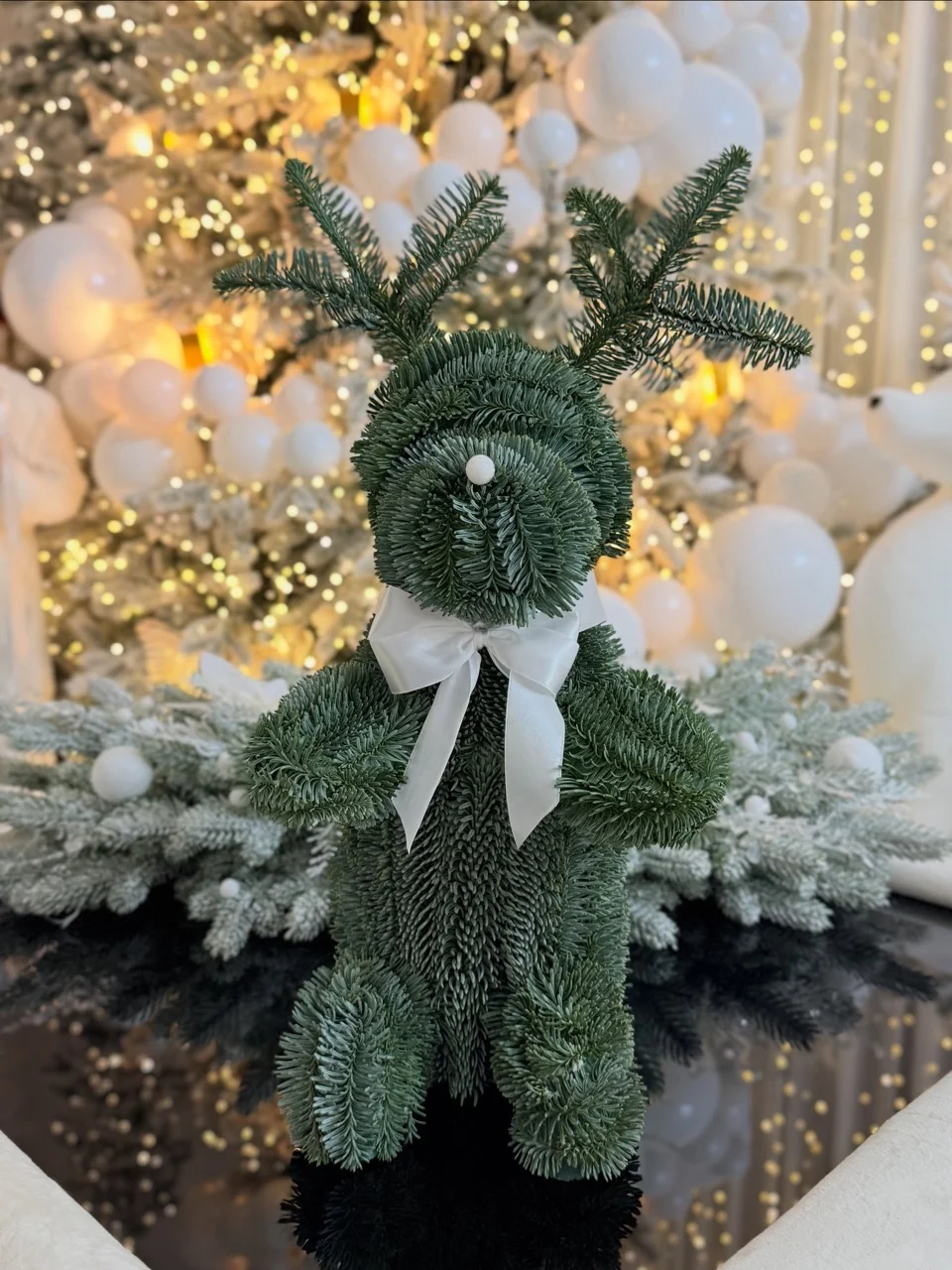 Green foliage reindeer decoration with a white bow in front of a softly focused white-and-gold Christmas tree.