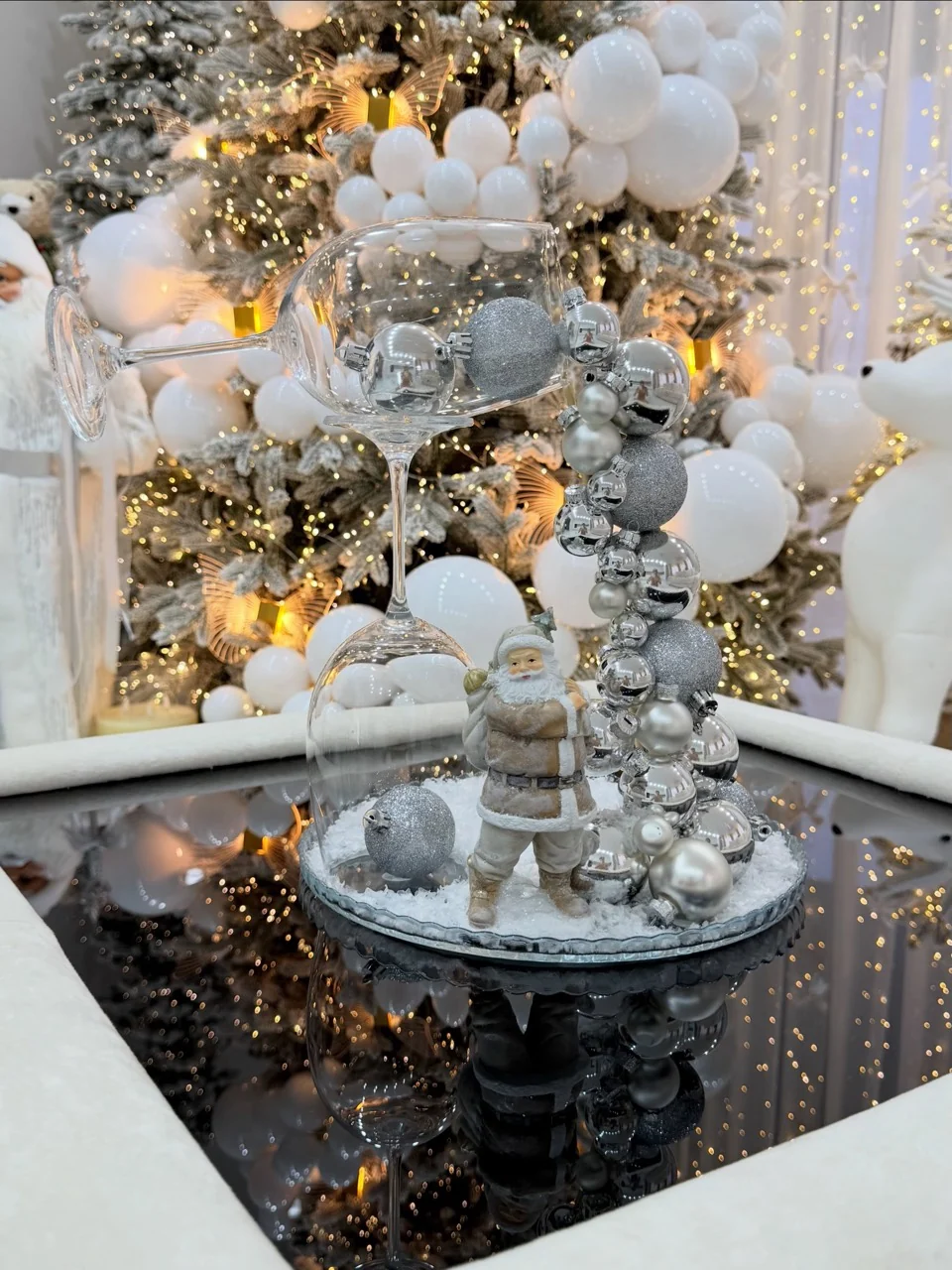 Glass and silver-bauble tabletop decoration with a small Santa figure on a mirrored table in front of a white Christmas tree.