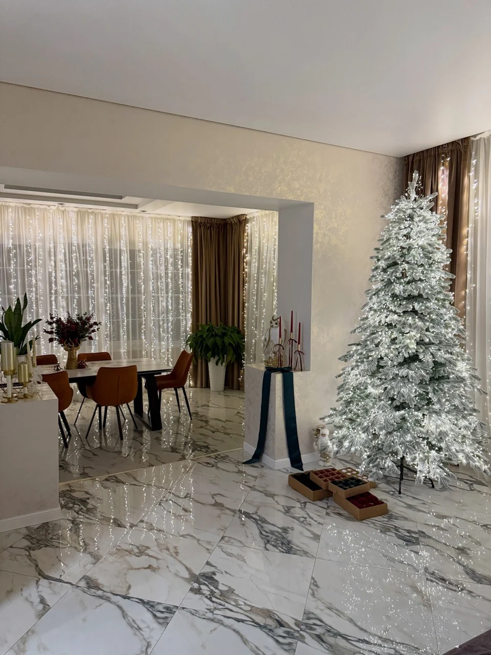 Living and dining room with curtain lights and an illuminated flocked Christmas tree during ornament preparation.