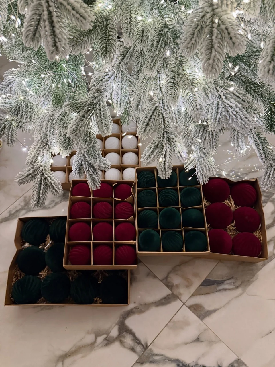 Boxes of burgundy, emerald and white baubles beneath illuminated snow-covered tree branches.