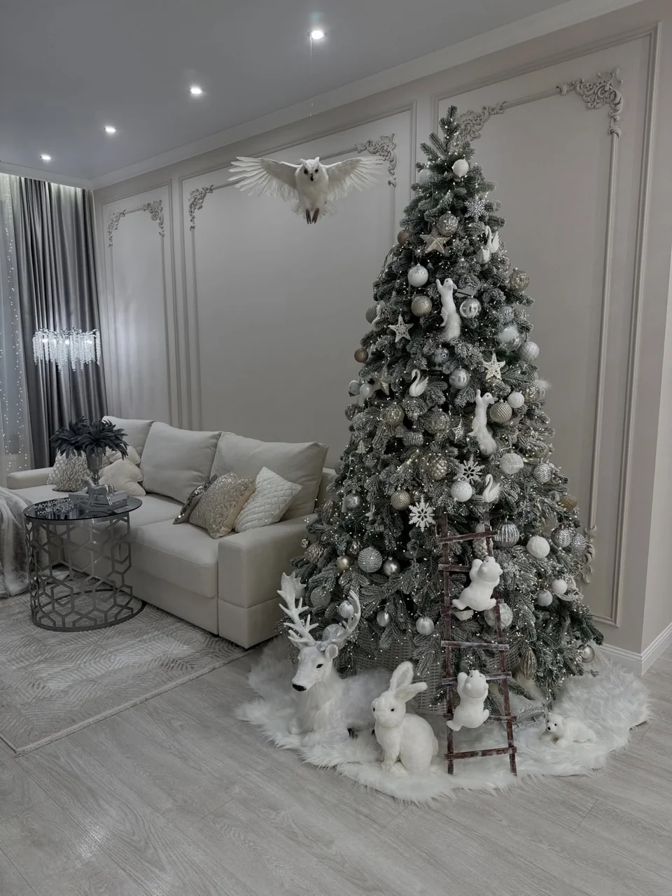 Christmas tree with silver and white ornaments, woodland animal decorations and a suspended white owl beside a cream sofa.