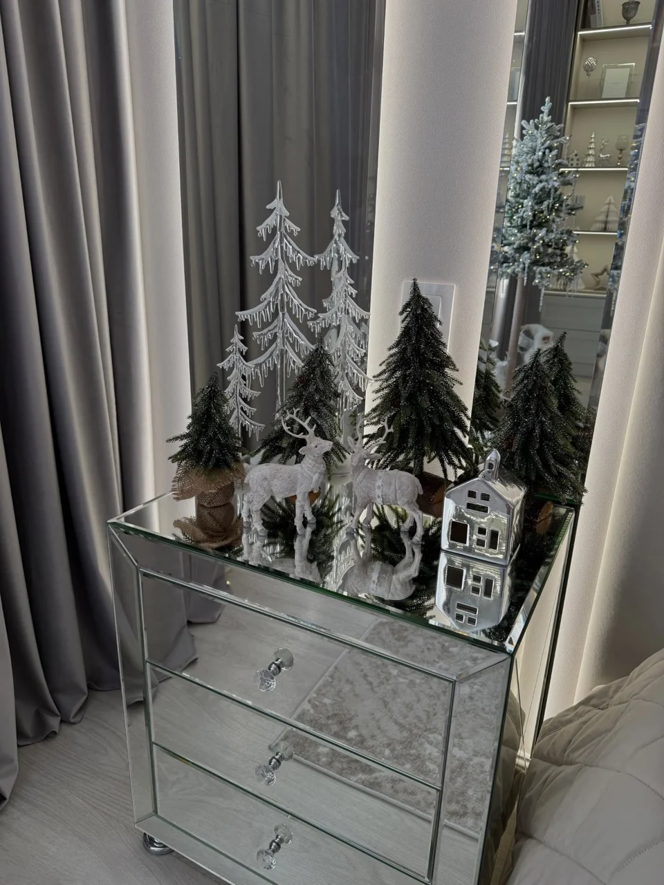 Miniature evergreen trees, white reindeer and a silver house arranged on a mirrored bedside cabinet.