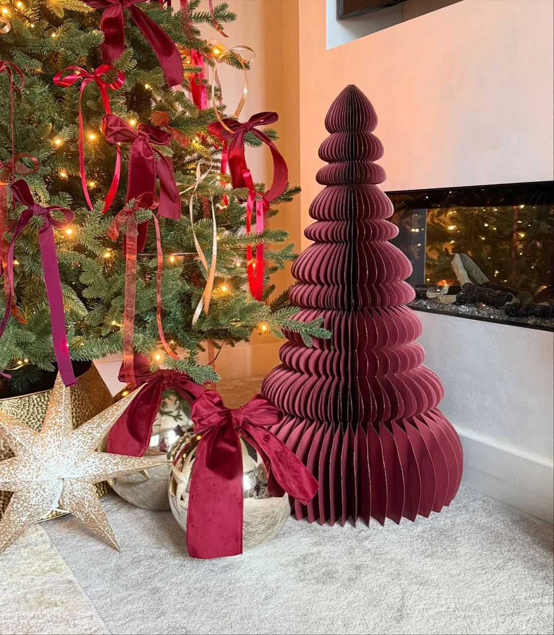Tall burgundy honeycomb paper tree beside a silver bauble with a burgundy bow and a gold star near a fireplace.