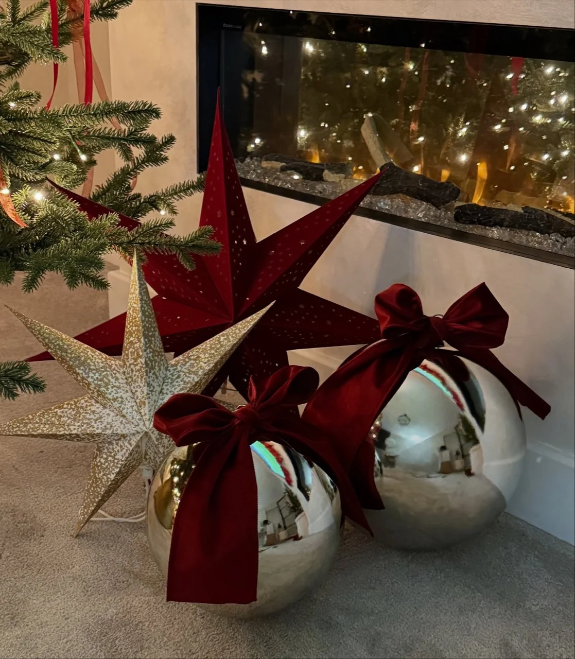 Reflective metallic baubles tied with burgundy velvet bows beside red and gold stars in front of a fireplace.