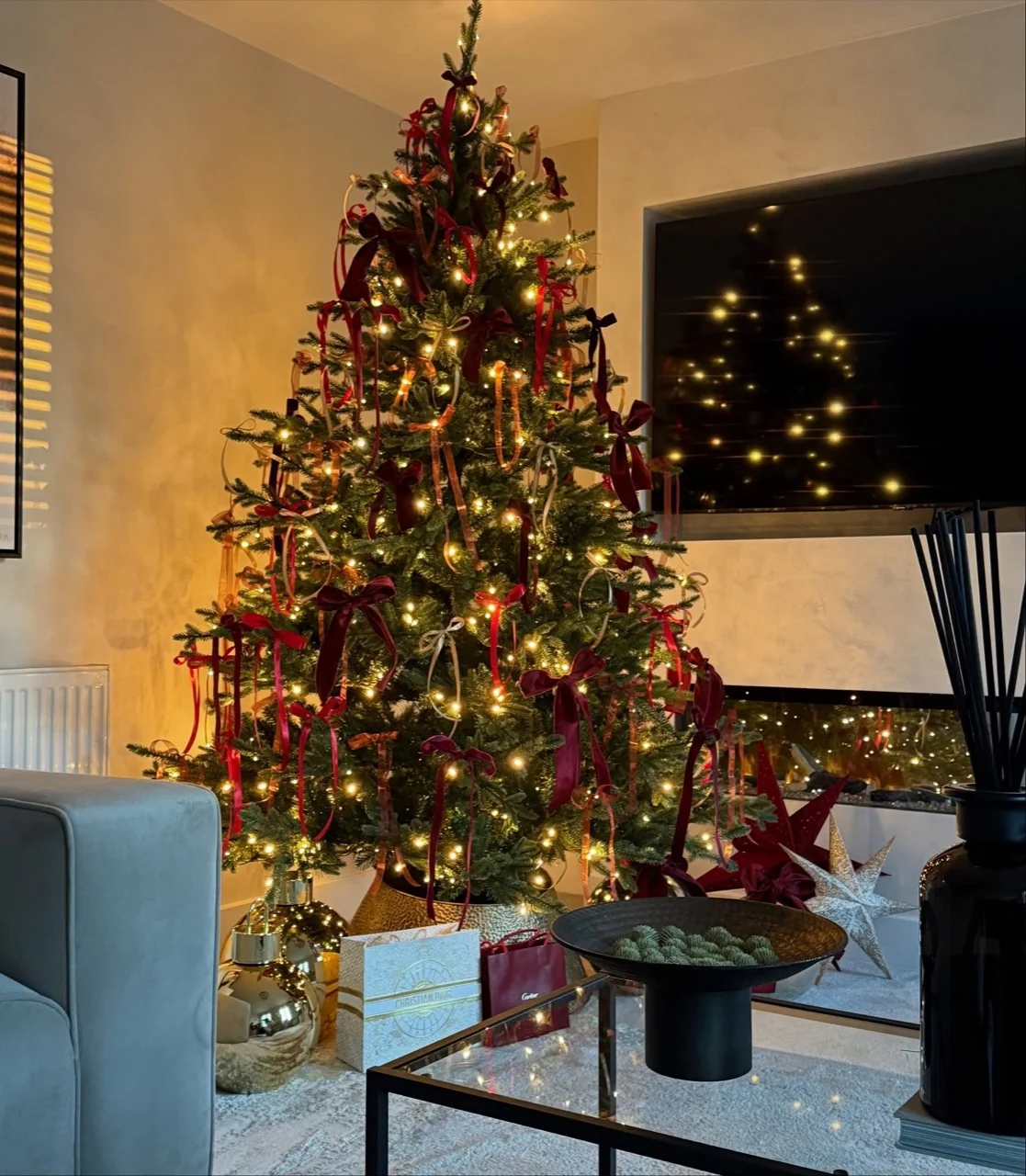 Christmas tree with trailing burgundy ribbons and warm lights beside a fireplace and television in a softly lit living room.