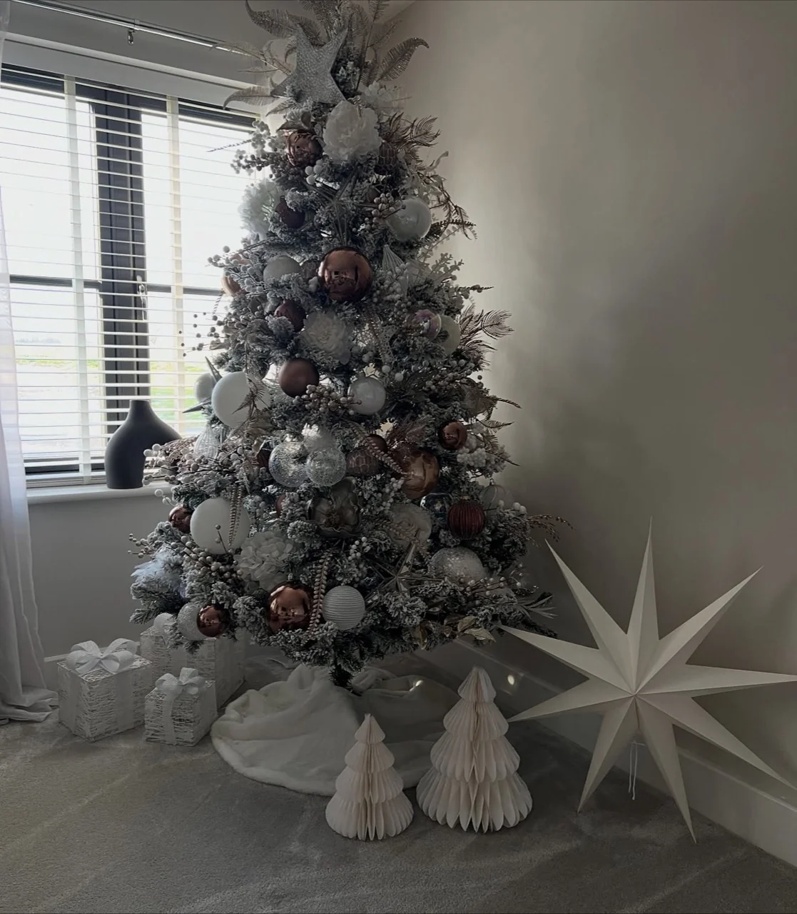Flocked Christmas tree with bronze and white ornaments beside wrapped white gifts, paper trees and a large white star.