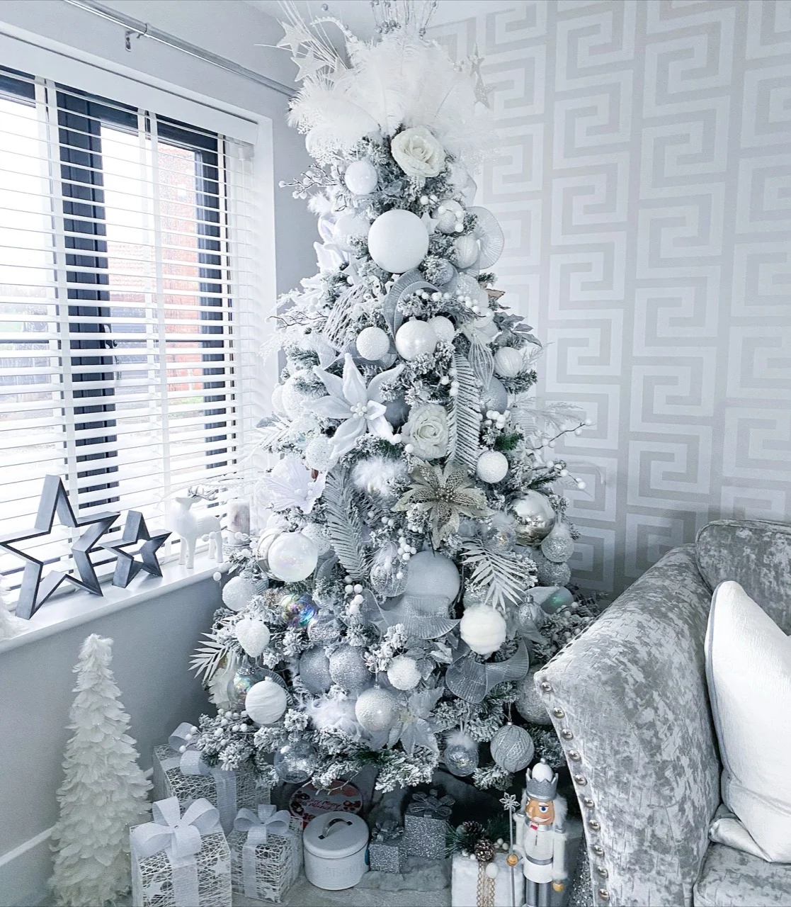 White and silver Christmas tree with a feather topper, flowers, berries and ribbon beside a grey sofa and window.